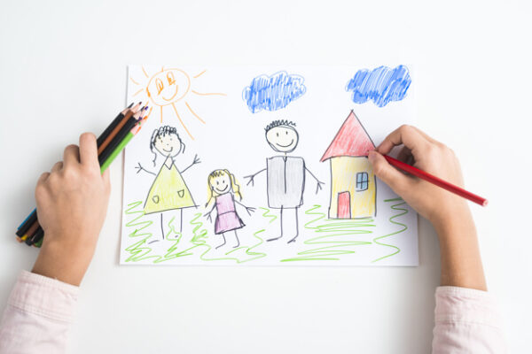 Criança desenhando a própria família com lápis de cor: casa, sol, pai, mãe e filha.